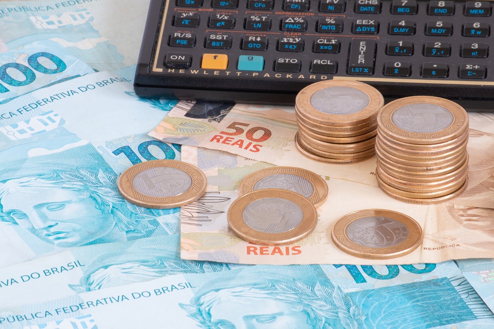 notas de cem e cinquenta reais espalhas na mesa, em cima tem moedas de um real e ao lado uma calculadora simulando qual o banco tem a menor taxa de juros para empréstimo
