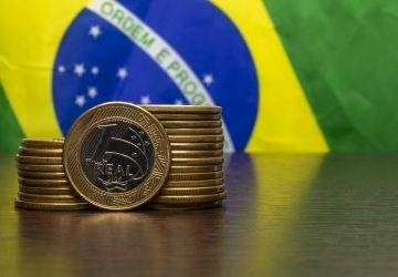 bandeira do Brasil ao fundo e a frente moedas de um real empilhadas, demonstrando o que muda com o novo aumento do salário mínimo em 2022