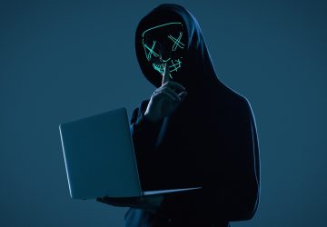 Homem anônimo em um capuz preto e máscara de néon invadindo um computador, simulando os golpes com jogos NFT